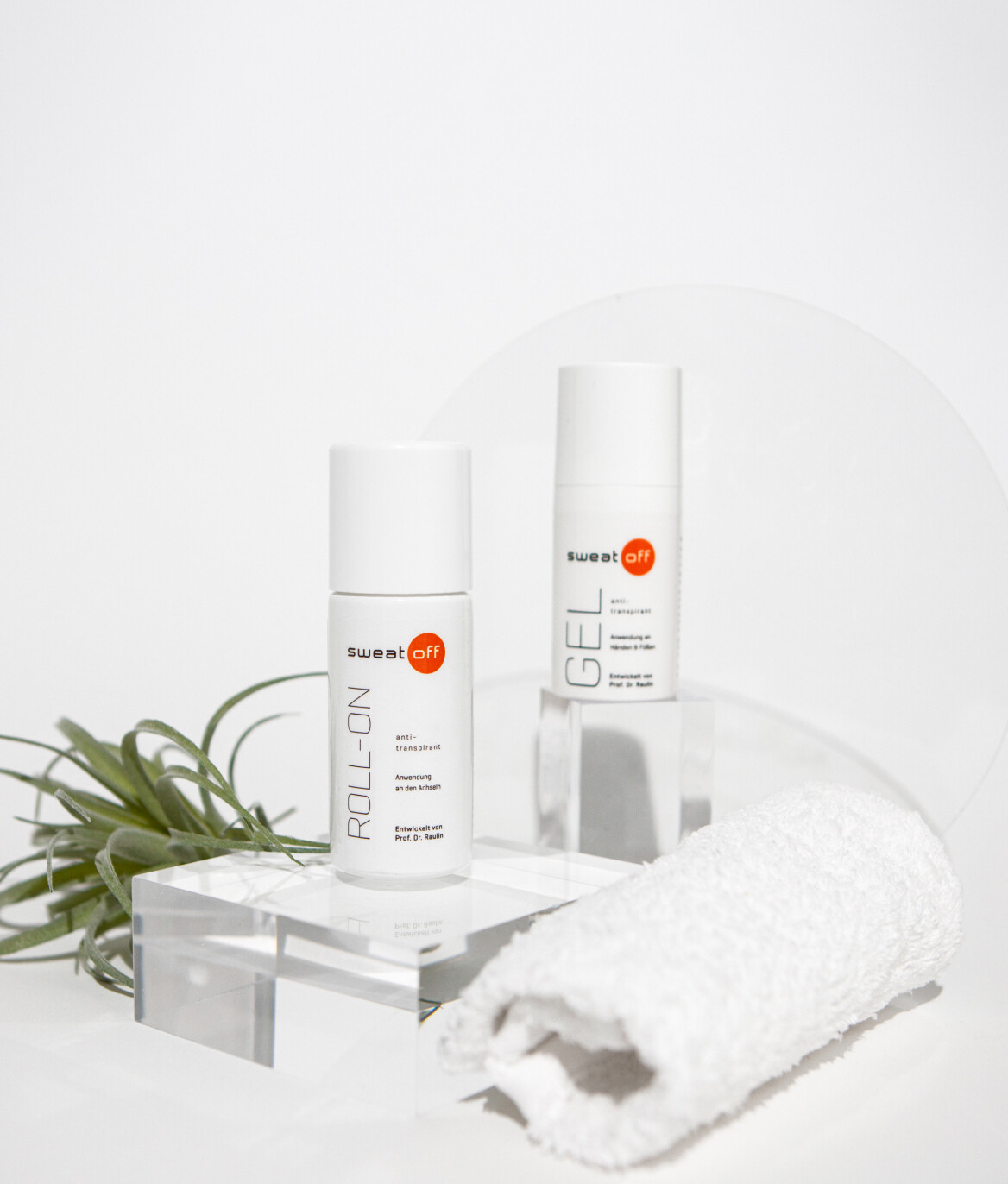 Roll-On und Gel platziert auf transparentem Podest. Handtuch zusammengerollt rechts davor, Pflanze links dahinter